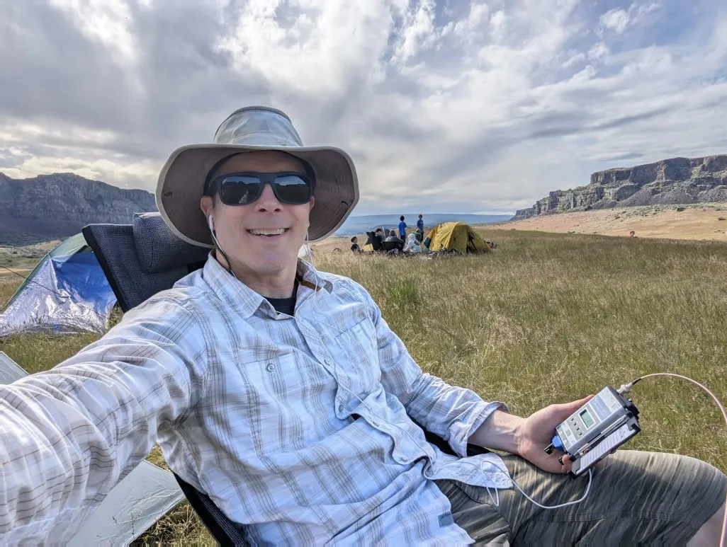 Justin operating a portable radio at a campsite in an open valley beneath basalt cliffs.