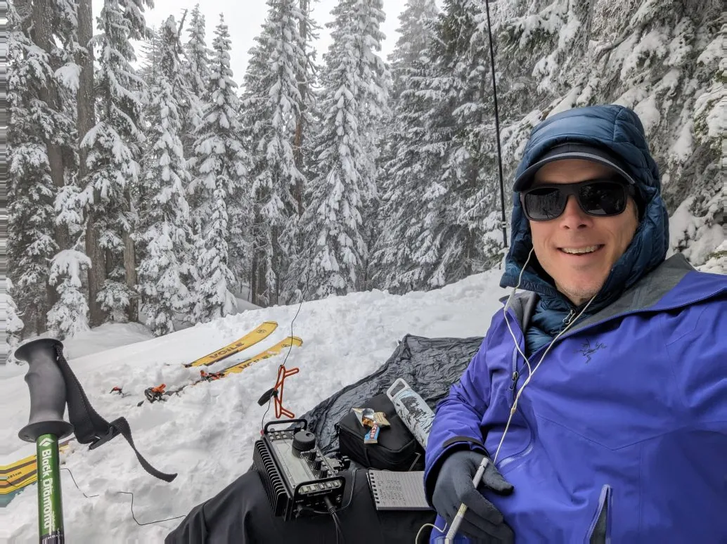 Justin with a portable radio and skis in a snow-covered forest.